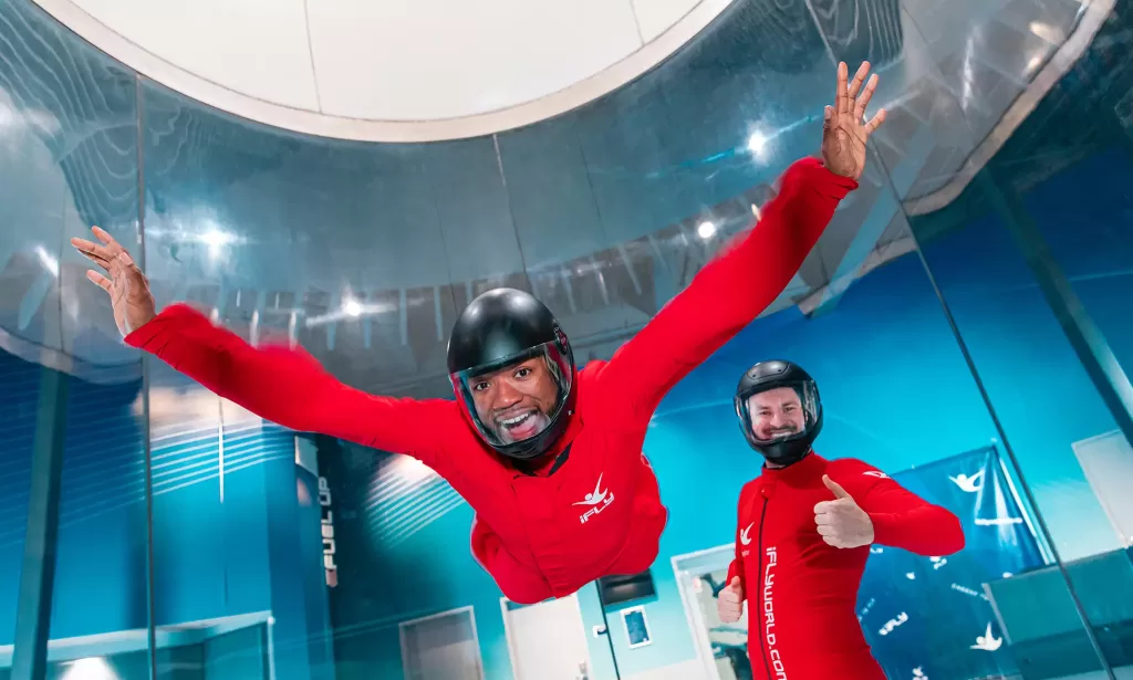 Participant enjoying iFLY indoor skydiving in a wind tunnel wearing a red flight suit and helmet with a thumbs-up from the instructor in the background.